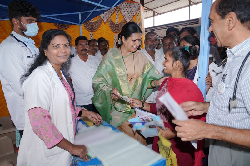 A woman in a green silk saree hands a health card to a smiling young woman at an SS Care Trust Spandana registration desk, with doctors and visitors around them.