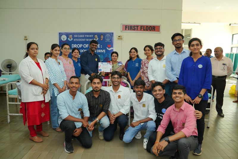 A large group of Student Wing volunteers and staff pose indoors after a blood donation drive, with a certificate on display.