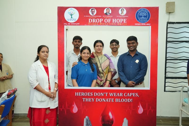 Five people pose inside a red photo-booth frame for a blood donation drive, with a staff member standing beside it.