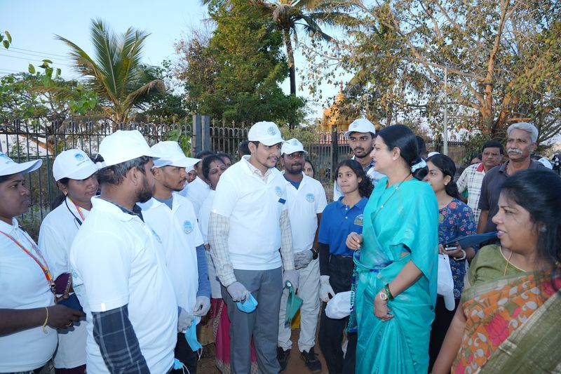 Volunteers in white caps gather outdoors with a trustee during a community awareness drive near a park gate.