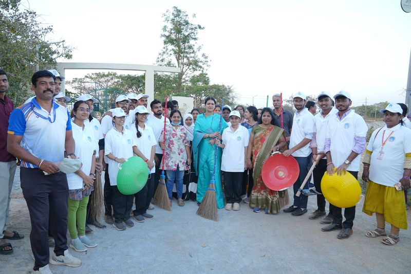 Volunteers in white caps hold brooms and colourful baskets for a community cleanliness drive, with a trustee among them.