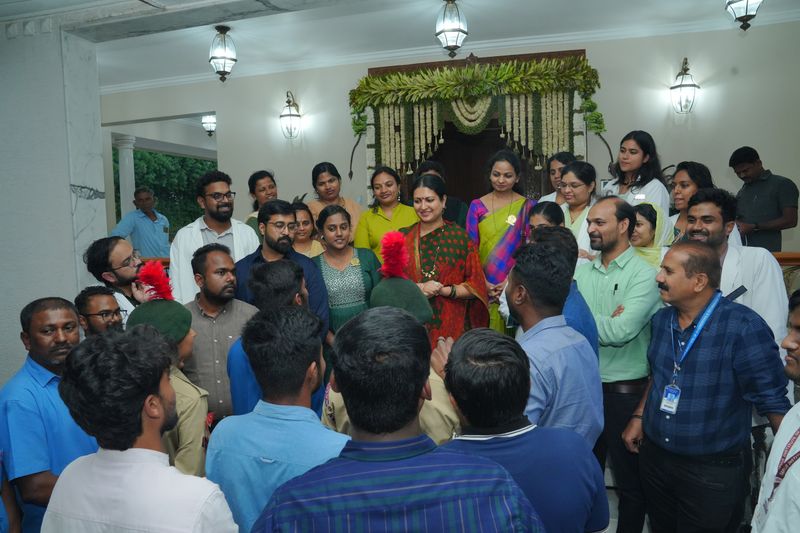 A crowd gathers indoors around a trustee near a flower-garlanded doorway during a Trust function.