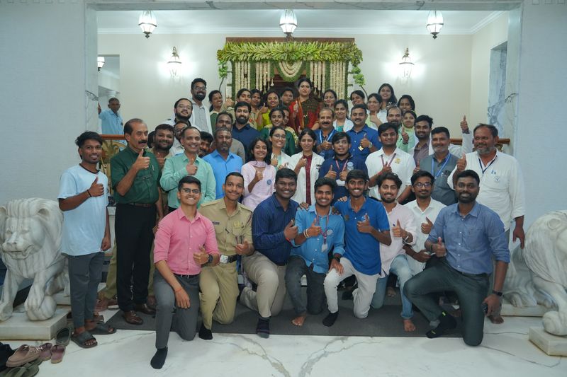 A large group of student volunteers and staff pose together indoors giving a thumbs up, with stone lion statues nearby.