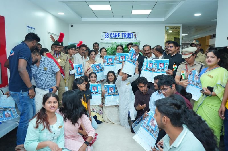 A crowd including a scout in uniform holds up printed calendars with photographs, standing beneath an SS Care Trust sign.