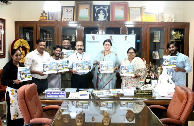A group poses in an office holding up calendars with photographs, in front of a cabinet of awards and mementos.