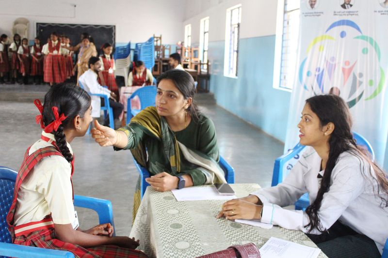 A health worker gently checks a schoolgirl's face while a colleague reviews papers at a child health screening camp.