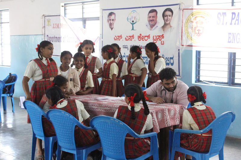 A group of schoolchildren in uniform gather around a health worker seated at a table during a school health check-up.
