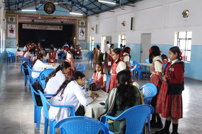 A wide view of a school hall with students and health workers seated at tables during a child health screening drive.