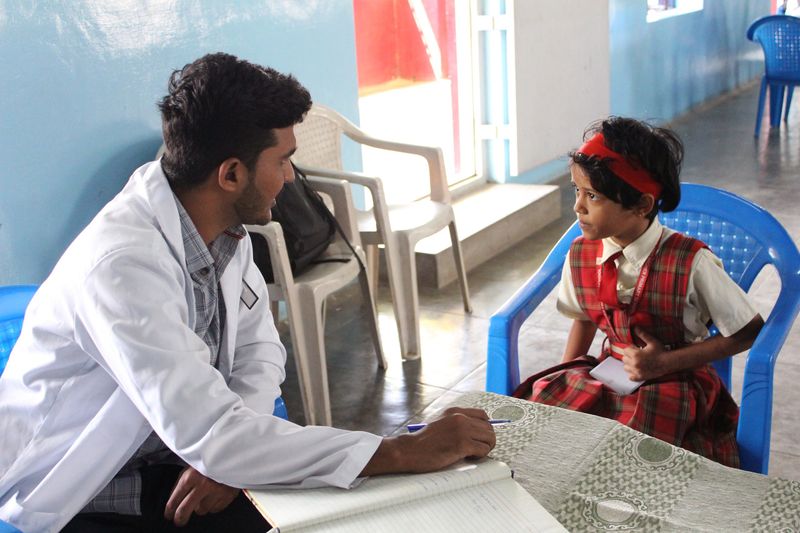 A male health worker in a white coat talks with a young girl seated across a table during a health check-up.