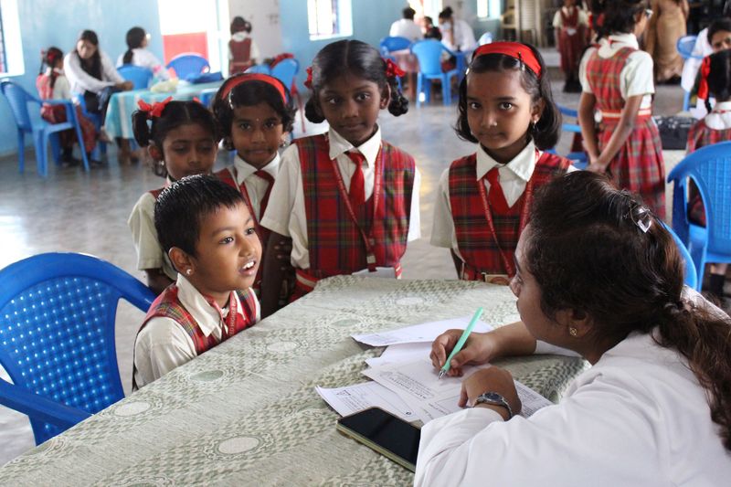 A group of young children stand around a table as a health worker takes notes during a school health check-up.