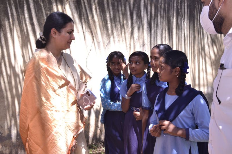 A woman in an orange saree talks with four schoolgirls in blue-and-white uniforms beside a sunlit wall.