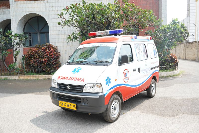 An SS Care Trust Maruti Eeco ambulance with a 24x7 emergency service sticker, parked outside a hospital building.