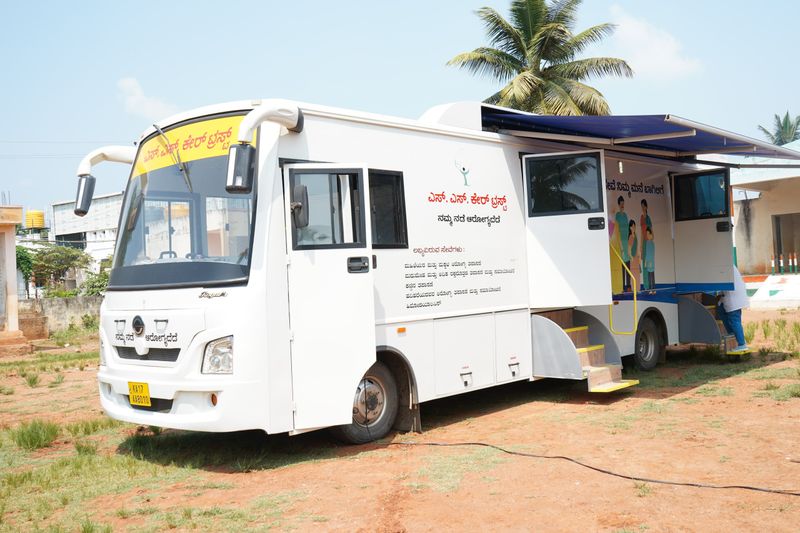 The SS Care Trust mobile health-camp bus parked at a village camp, its side awning open and steps down.