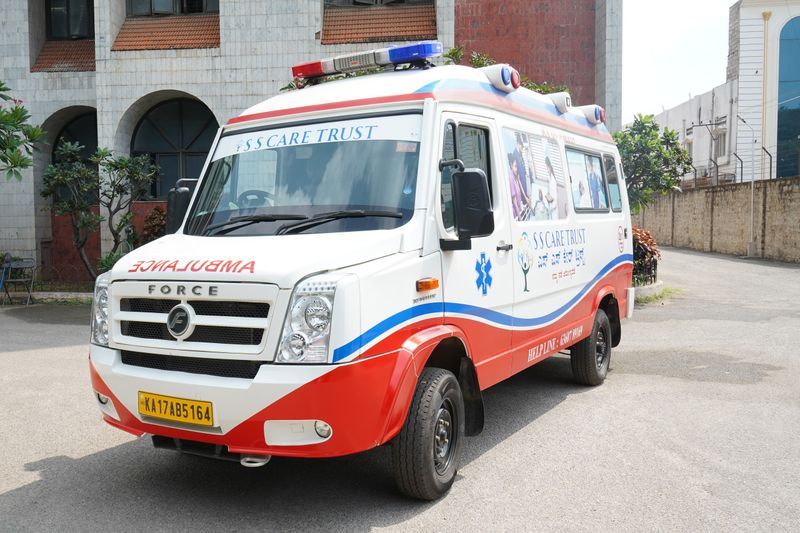 An SS Care Trust Force Traveller ambulance with the Trust's helpline painted on its side, parked outside a hospital building.