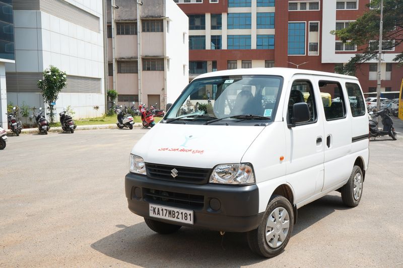 A white SS Care Trust Maruti Eeco van with the Trust's name on the bonnet, parked on a college campus.
